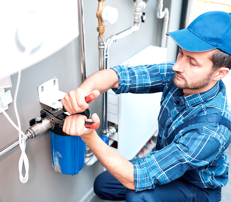 Young man in workwear using pliers while installing water filtration system in the kitchen of client